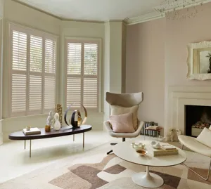 Neutral living room with modern cream chairs, round low table on a abstract patterned rug in shades of cream and brown, wooden low coffee table sits in the alcove of a bay window dressed in cream shutters.