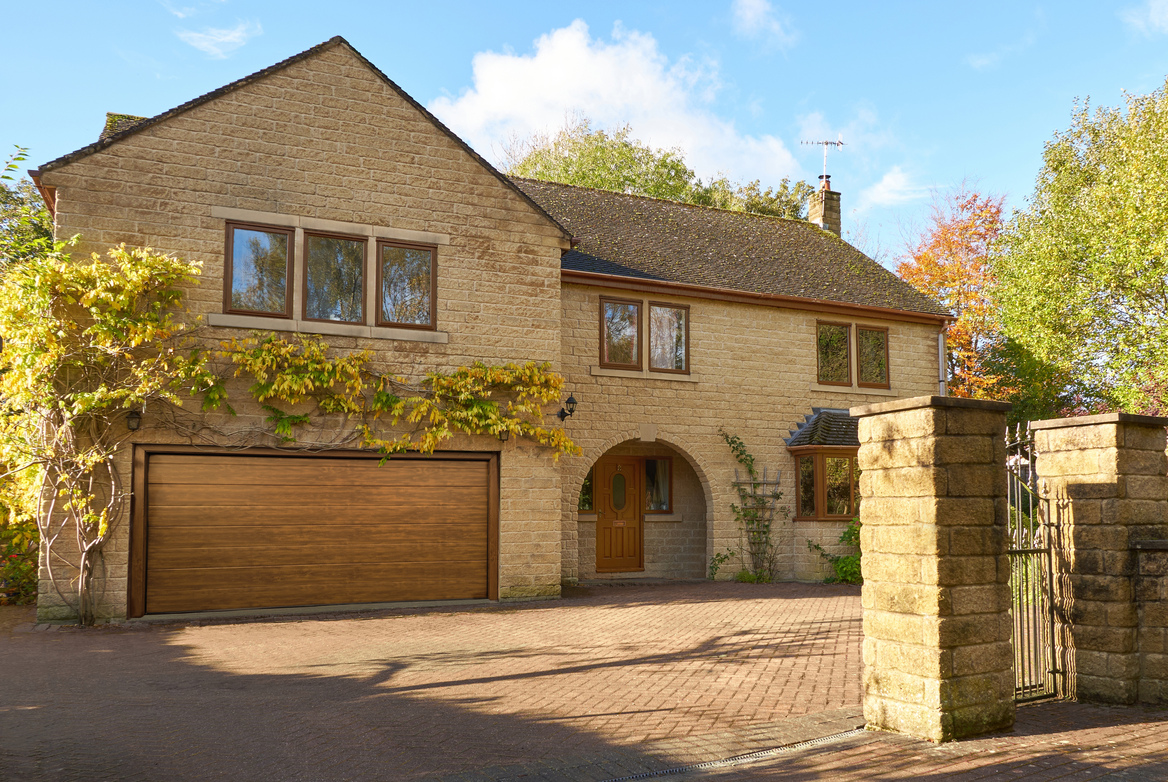 Large Yorkshire-style stone house and drive with integral garage door in mid brown matching front door, with climbing plant above garage.