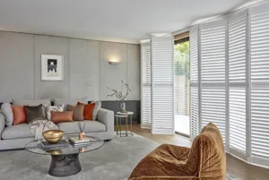 Grey living room with wooden floor and grey rug, grey sofa with cushions in browns and coppers, glass circular coffee table, copper soft chair and floor to ceiling windows and doors in white tracked shutters, partially open.