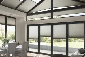 Open plan lounge diner glass gable end extension with white wood table and chairs, sofa with throws, and grey Duette blinds to doors and windows and angled roof windows.