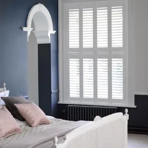 Bedroom with dark blue walls, white bed with grey and dusky pink cushions, grey upholstered stool at bed, archway with white moulding and on right window dressed in white tier on tier shutters, closed with louvres open.