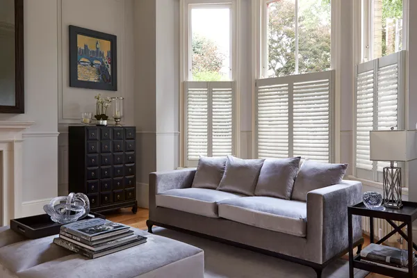 Cream lounge with bay window dressed in white birch cafe style shutters, closed with louvres open, silver grey sofa and large footstool as table, wooden side table and dark wood apothecary cabinet.
