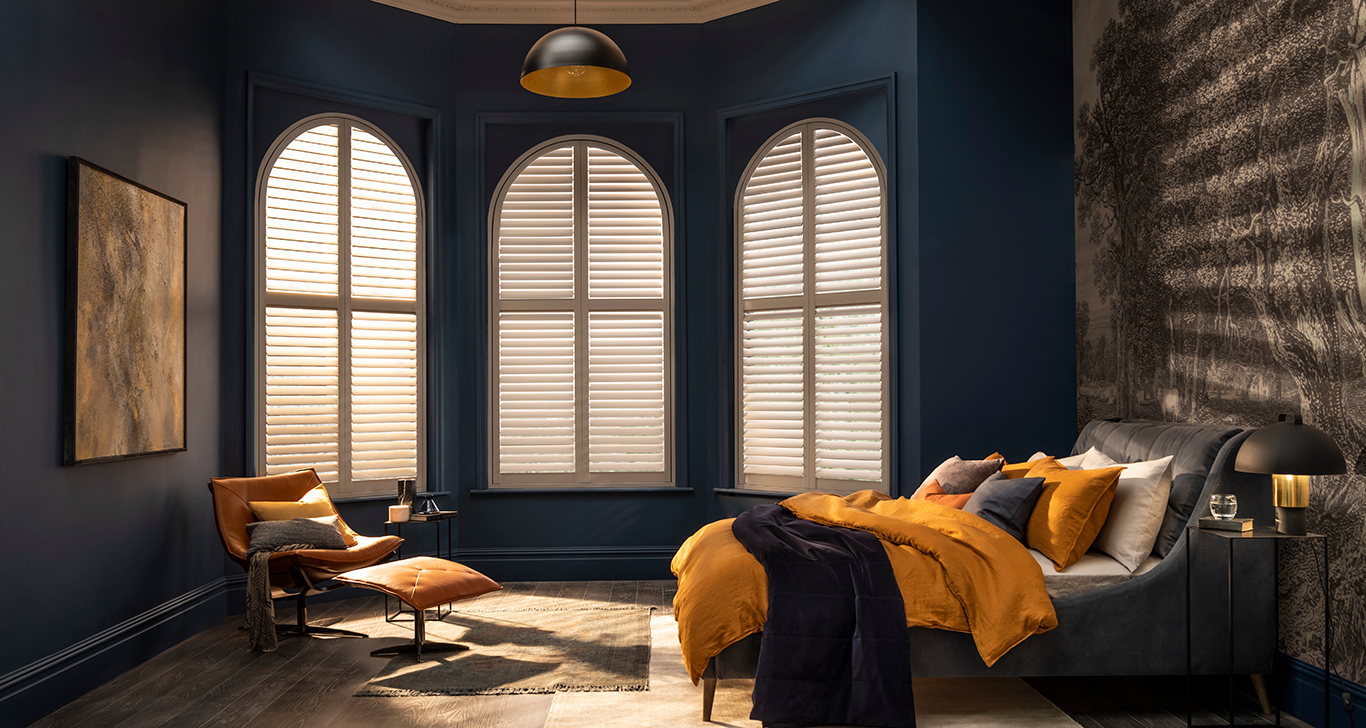 Bedroom with dark blue walls left and rear, brown striped fabric wallcovering on right wall behind bed dressed in copper and navy blue bedding, to rear three large arched windows in a bay, dressed with white shutters, closed with louvres open.