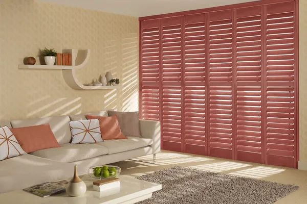 Living room in pale corn yellow with tall window dressed in full height shutters in salmon with sofa in pale peach with contrasting peach and salmon cushions.