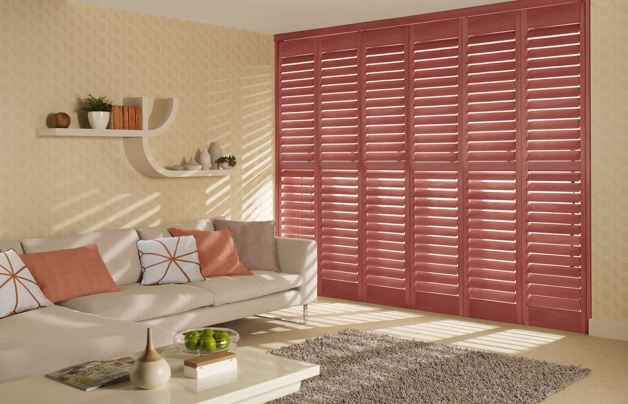 Living room in pale corn yellow with tall window dressed in full height shutters in salmon with sofa in pale peach with contrasting peach and salmon cushions.