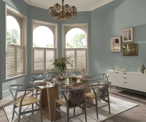 Dining room with three windows on a curved wall dressed in white cafe style shutters, closed with louvres open, pale blue walls, modern glass dining table with wood and metal chairs, white low sideboard.