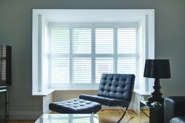 Lounge with inset bay window dressed in white shutters, closed with louvres open, dark brown leather lounger chair in recess, glass coffee and side tables, dark brown leather sofa and black side lamp.