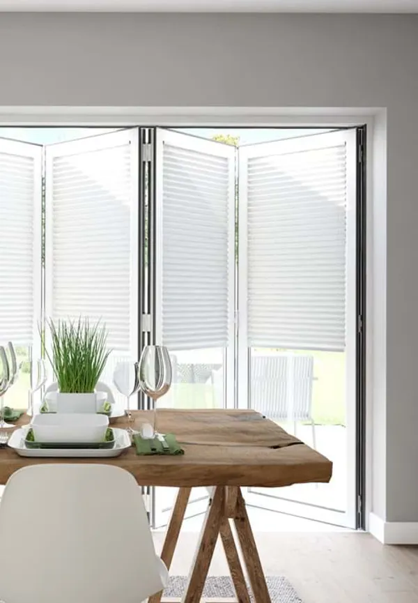 Dining room in pale grey with pale wood floor, wooden trestle-style table with white contemporary chairs, table set with white crockery and bifold doors dressed with white blinds within frame, doors partially open.