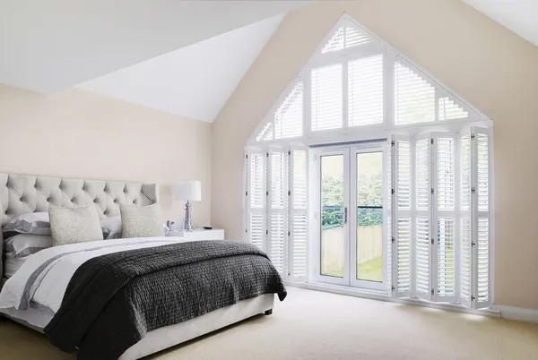Bedroom with pale pink walls, bed wth quilted headboard dressed in white and grey bedding, gable end window and doors dressed in white shutters, doors open.