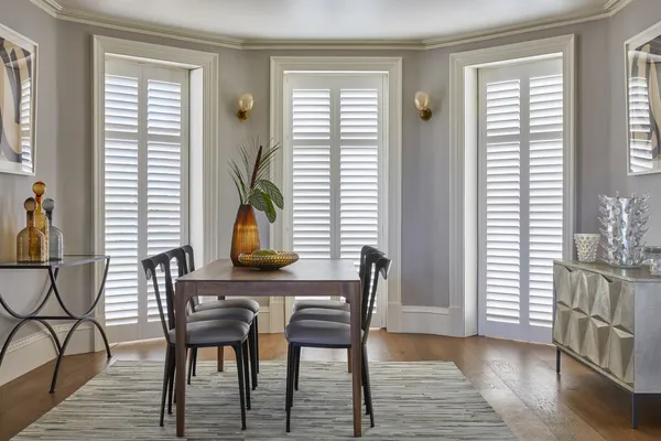 Dining room with wood floor,  three floor to ceiling windows in an extended bay dressed in white shutters, closed with louvres open, rug in grey tones, wooden table and chairs with grey cushioned seats, amber vase and greenery on table.