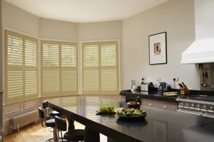 Kitchen with cream walls, black gloss surfaces including an island with black high-back stools and a bow bay window dressed in cream shutters, closed with louvres open.
