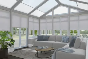 Rectangular conservatory with dark wood floor, pale grey L-shaped sofa, large rug and leafy plant in pot by door with pale lilac roller blinds on side windows and off-white pleated blinds in roof.