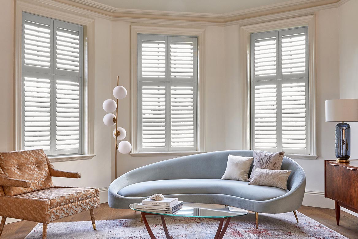 In a living room that is decorated in white are Sketched Harlequin Shutters which are fitted to bay shaped windows in a room with an armchair, curved grey sofa and a glass coffee table 