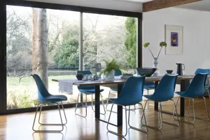 Dining room with wood floor, modern table and blue chairs and full sized patio doors with view out over countryside garden.