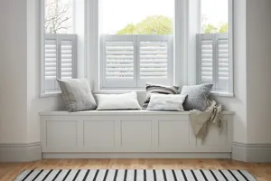 White room with pale wood floor, white rug with blue stripes in front of a bay window with a window seat filled with white and pale grey cushions and white cafe style shutters, closed with louvres open.