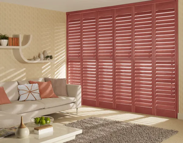 Living room in pale corn yellow with tall window dressed in full height shutters in salmon with sofa in pale peach with contrasting peach and salmon cushions.
