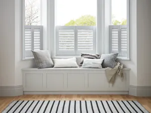 White room with wooden floor, white and blue striped rug in front of a window seat with pale blue and white cushions and cafe style shutters to window, in white.