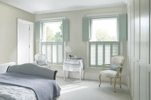 Bedroom with two adjacent windows dressed in pale green tier on tier shutters, top open, louvres closed, bottom closed louvres open, Queen Anne style chairs and dressing table, pale olive walls, cream carpet bed with pale green bedding and pale blue waffle throw.