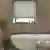 Bathroom with deep recessed window dressed in pale grey-beige roller blind, walls with wood panelling, roll top bath with central mixer taps, small bay tree in pot to left.
