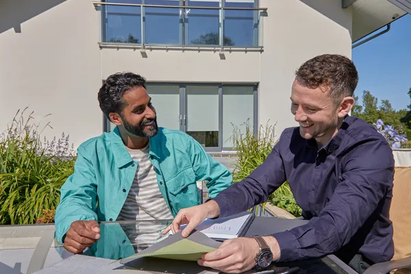 Exterior of modern house, two men looking at awning samples, seated at a table.