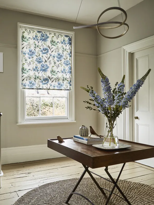 Hallway with wooden floor with round jute rug under folding card table with vase of blue flowers, white stairs to left and window dressed in roller blind with large blue flowers and green foliage