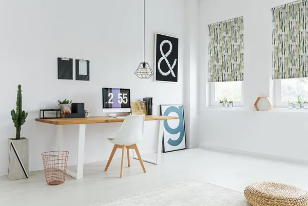 Large home office in white with  two windows dressed in roller blinds in pale green, black and yellow waterfall pattern, modern wood desk and chair, iMac and black desk accessories, tall cactus in concrete pot on floor by copper wirework bin.