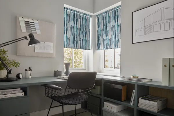 Home office corner with two narrow corner windows dressed in teal and white impressionist-style roller blinds, neutral walls teal desk and low shelving on both walls with framed drawing plans and paint swatches to walls.