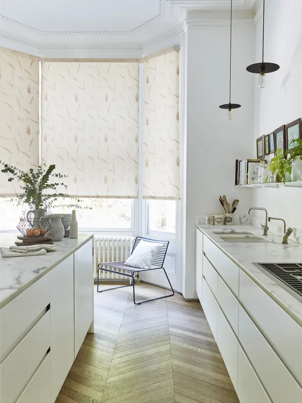 Kitchen in white with cream units and pale parquet floor, marble worktops and bat window dressed in roller blind in cream with wheatsheaf pattern.