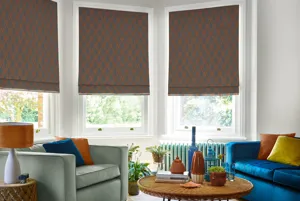 Modern lounge with blue sofa and grey chair with cushions in cobalt, copper and mustard, round coffee table with iron legs, bay windows dressed in three roman blinds in a greeny-grey with copper diamond and romboid pattern.