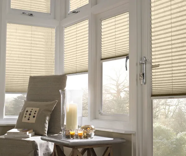 Corner of conservatory with upright chair with natural linen cover and cushion, small table with hurricane lamp and candle and small candles in jars, with windows and doors dressed in ivory pleated blinds at varying heights.