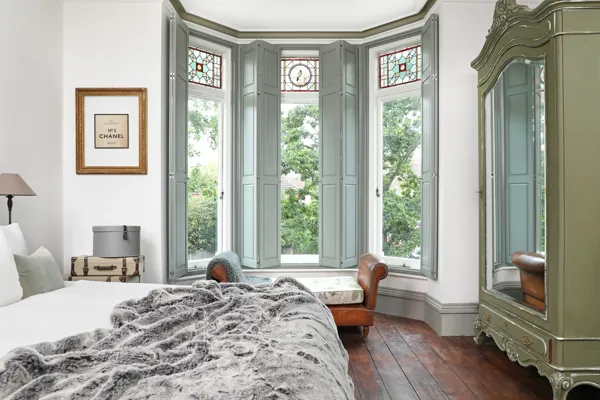 Classic-style bedroom with white walls, mid-grey painted skirting and bay window moulding, dark wood floor, bed on left with white bedding and grey faux fur throw, sage green armoir on left and bay window with patterned glass to top dressed in mid grey solid shutters.