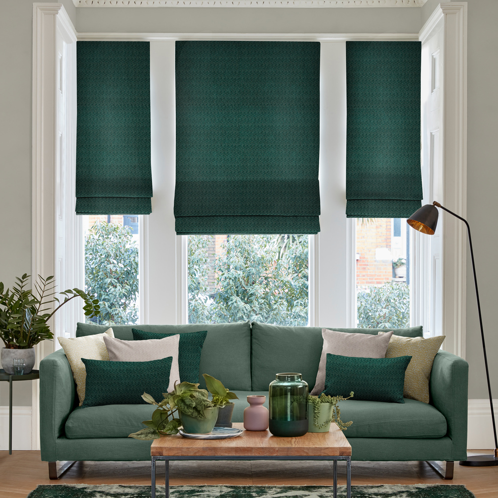 Pale grey lounge with bay window dressed in two narrow and one wide Roman blinds in deep green with pale wood floor and large faded geometric patterned forest green rug with table and green accessores, green sofa with cushions in dark green and cream.