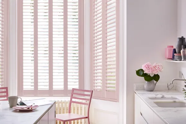 Close up of kitchen with bay window dressed in pale pink shutters, louvres open, white walls with grey marble worktop and island, pink wooden chairs, vase with pink flowers.