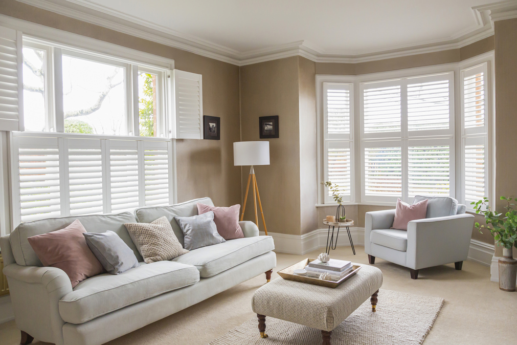 Neutral lounge with cream carpet and rug, pale grey sofa in front of window dressed with white tier on tier shutters, top half open, pale grey chair in a curved 5-bay window dressed in white shutters, louvres open.