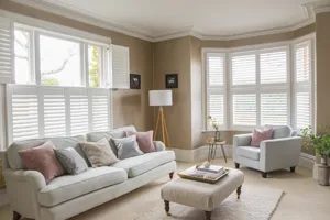 Neutral lounge with cream carpet and rug, pale grey sofa in front of window dressed with white tier on tier shutters, top half open, pale grey chair in a curved 5-bay window dressed in white shutters, louvres open.
