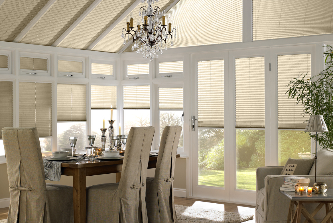 White conservatory with dining table and chairs dressed with beige linen covers, table set with glassware and candles, chandelier above and windows and roof dressed in ivory pleated blinds at various heights.