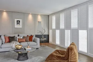 Neutral lounge with wood floor, grey rug, grey sofa and copper velvet beanbag-style chair with round glass pedestal coffee table and full length floor to ceiling windows and doors dressed in tier on tier white shutters, alternate louvres open.
