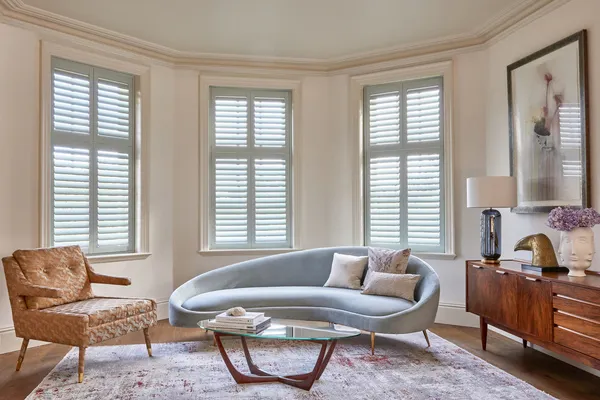 Lounge with curved bay windows dressed in pale Seaglass-coloured shutters, wooden chair on left upholstered in deep peach, pale blue shaped contemporary sofa in middle, wooden sideboard on right.
