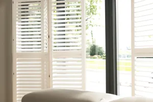 Close up in living room with brown sofa in front of patio doors dressed in white shutters, partially open.