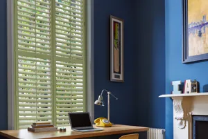 Home office with mid blue walls, white art deco style fireplace, pale wood desktop over black unit and tall narrow window dressed in pale green wooden shutters, closed with louvres open.