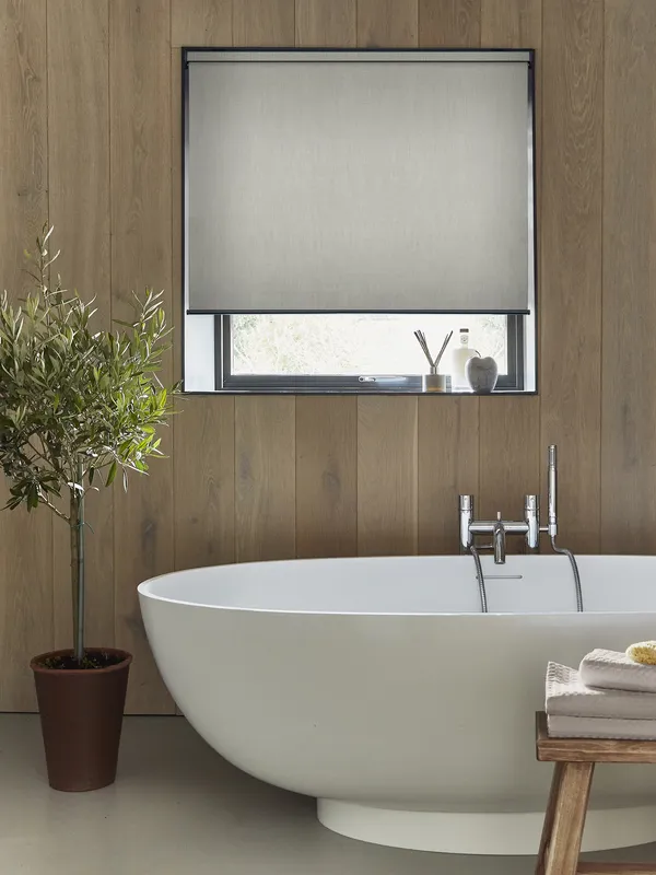 Bathroom with deep recessed window dressed in pale grey-beige roller blind, walls with wood panelling, roll top bath with central mixer taps, small bay tree in pot to left.