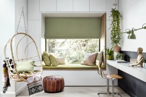 White room with hanging egg chair, white desk shelving with wood chair, deep recess window with green cushion seating dressed in an olive green Roman blind partially lowered.