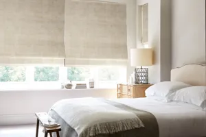 Neutral bedroom, bed with white bedding, grey/brown throw across bed, small cream wrap on top, pale wood beside table with cream lamp and large windows dressed in a Roman blind in ivory faux velvet on top of a white Duette blind.