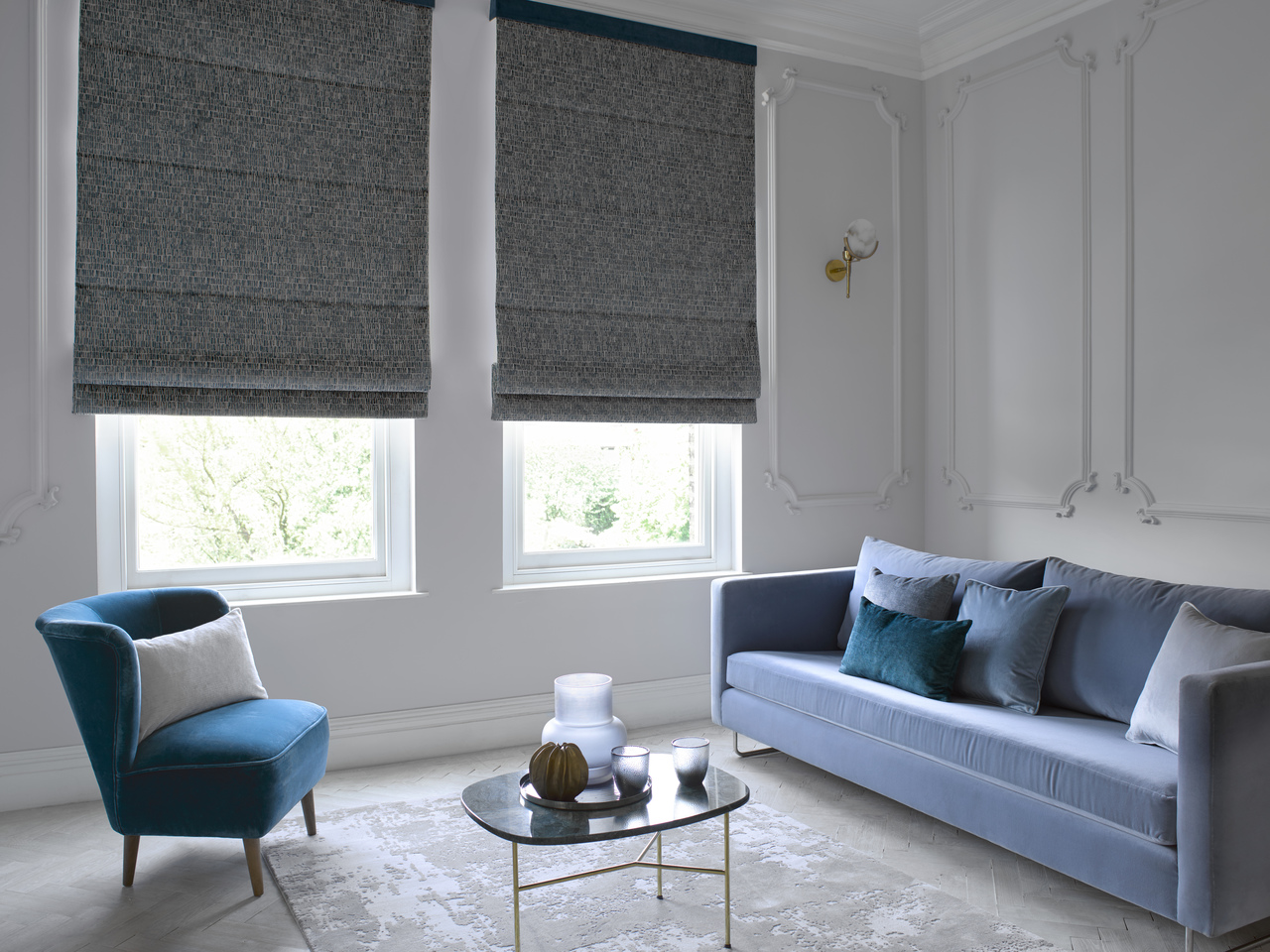 Pale grey living room with two adjacent tall windows dressed in dark grey Roman blinds three quarters lowered, mid blue sofa, cobalt blue chair and a small glass coffee table in between.