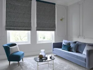 Pale grey living room with two adjacent tall windows dressed in dark grey Roman blinds three quarters lowered, mid blue sofa, cobalt blue chair and a small glass coffee table in between.