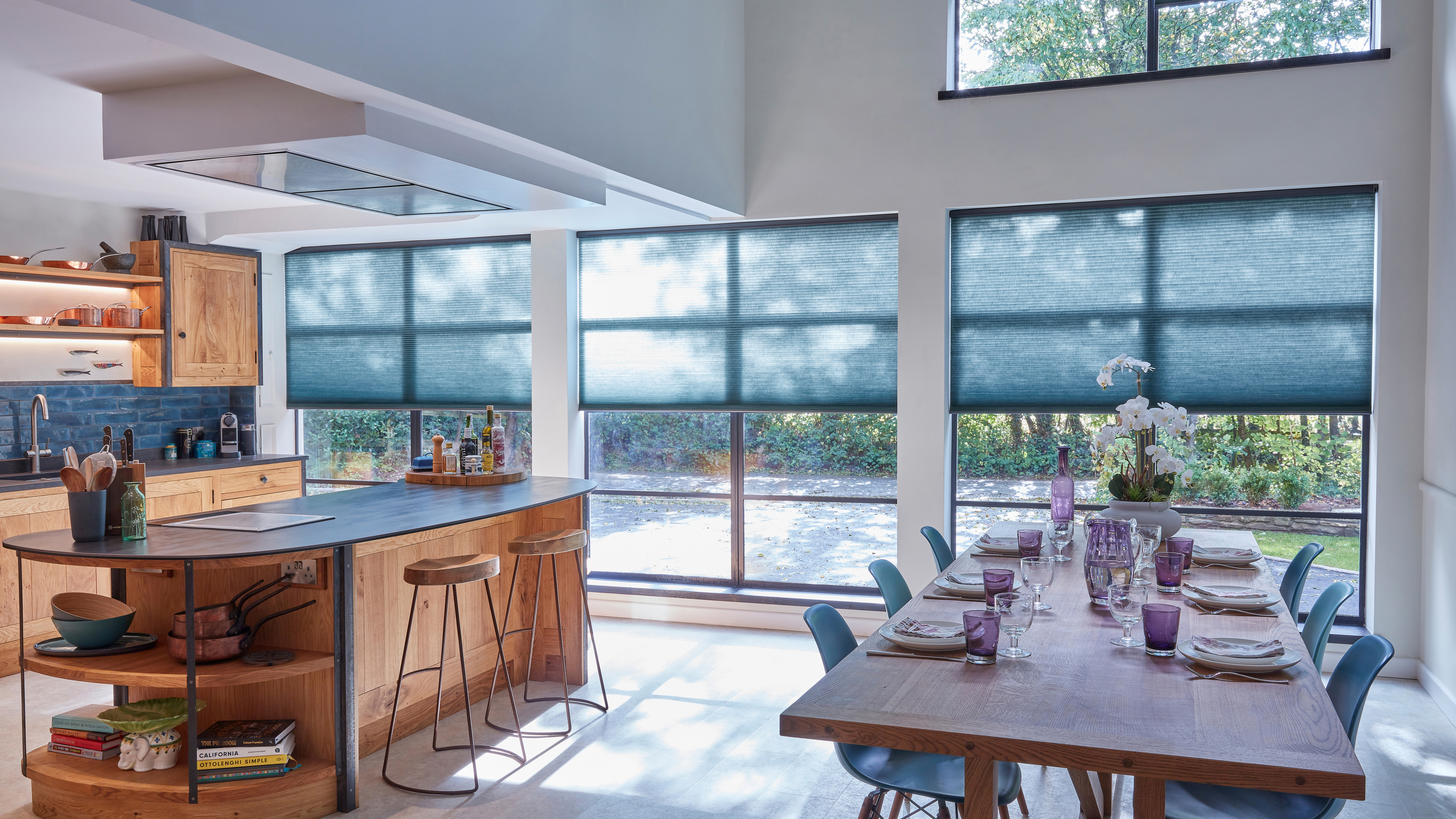 Open plan kitchen diner with windows dressed in mid blue duette blinds half raised, wooden units, wooden island and wooden table and blue chairs.