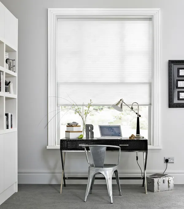 White pleated blind fitted to a tall rectangular window in astudy with grey walls, a metal chair and black desk 