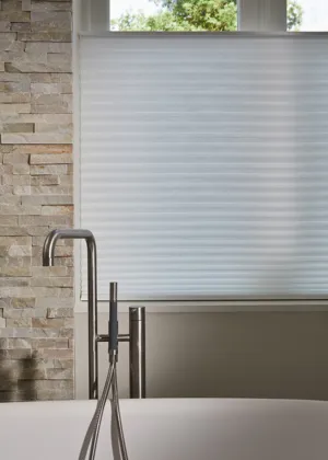 Close up of a window behind a bath with plain mixer tap, stonework walls and window dressed in white pleated blind raised from the bottom up.