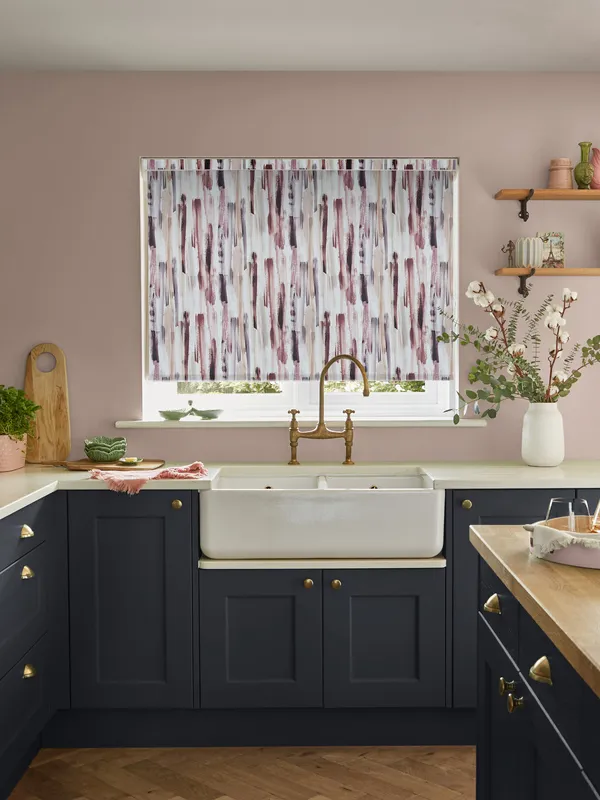 Kitchen with a window dressed with a roller blind in a pink, grey and beige waterfall pattern, dark blue cabinets, dusky pink walls and behind a butlers sink with antique brass mixer taps.