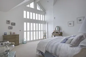Pale grey bedroom with bed in white and grey covers, upholstered grey bedhead and gable end windows and door dressed in white shutters, louvres open and tracked door shutters partially folded back.